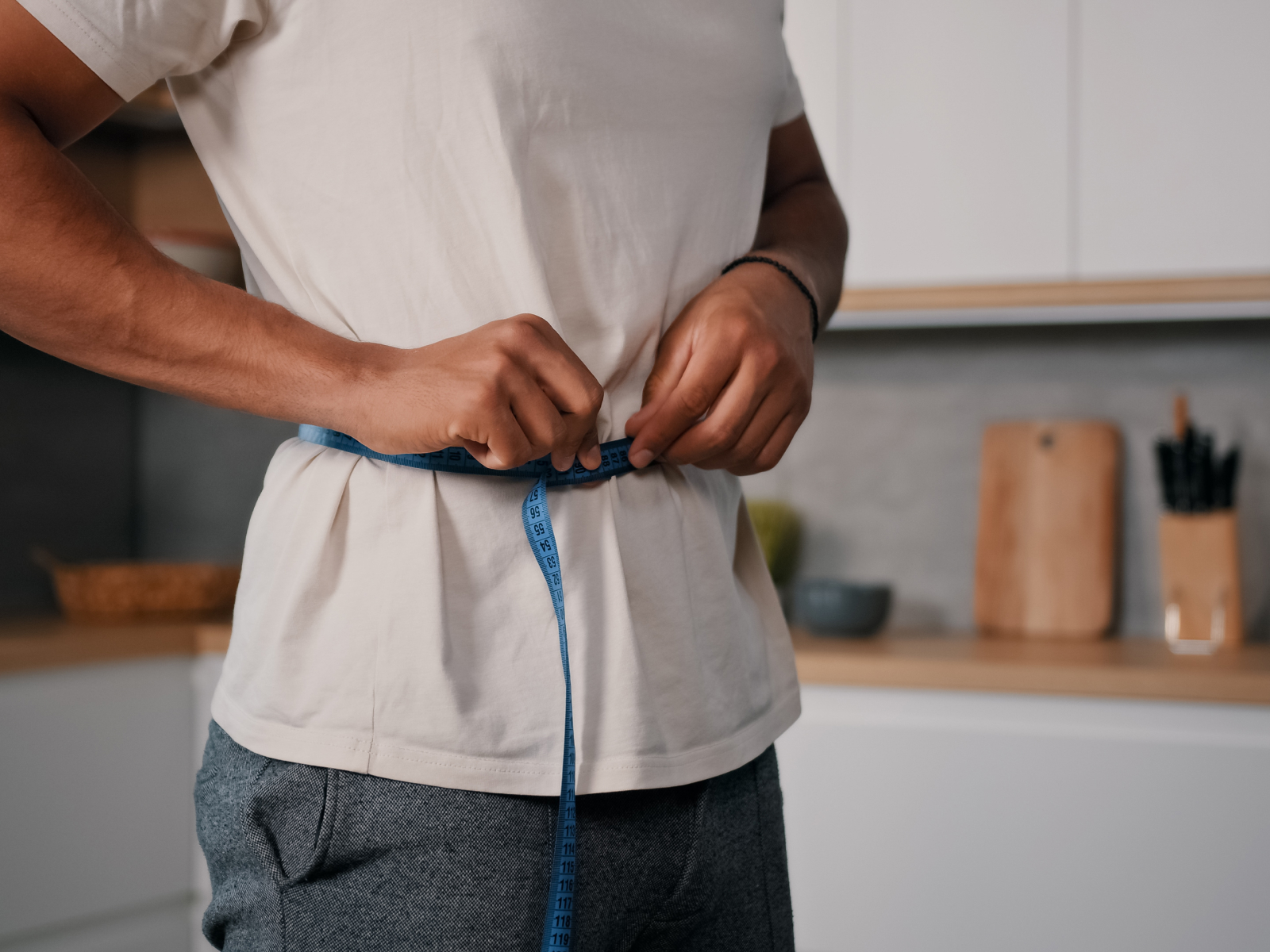 A man measuring his waist with a blue tape measure in a kitchen setting, symbolizing weight loss and appetite control; discover how tirzepatide suppress your appetite effectively.