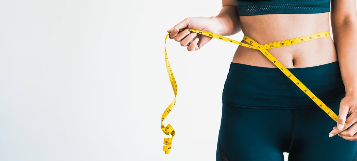 A fit woman in dark athletic wear wraps a yellow measuring tape around her waist, symbolizing weight loss and body transformation, highlighting the benefits of tesamorelin, which helps reduce abdominal fat, improve metabolism, and support overall wellness.