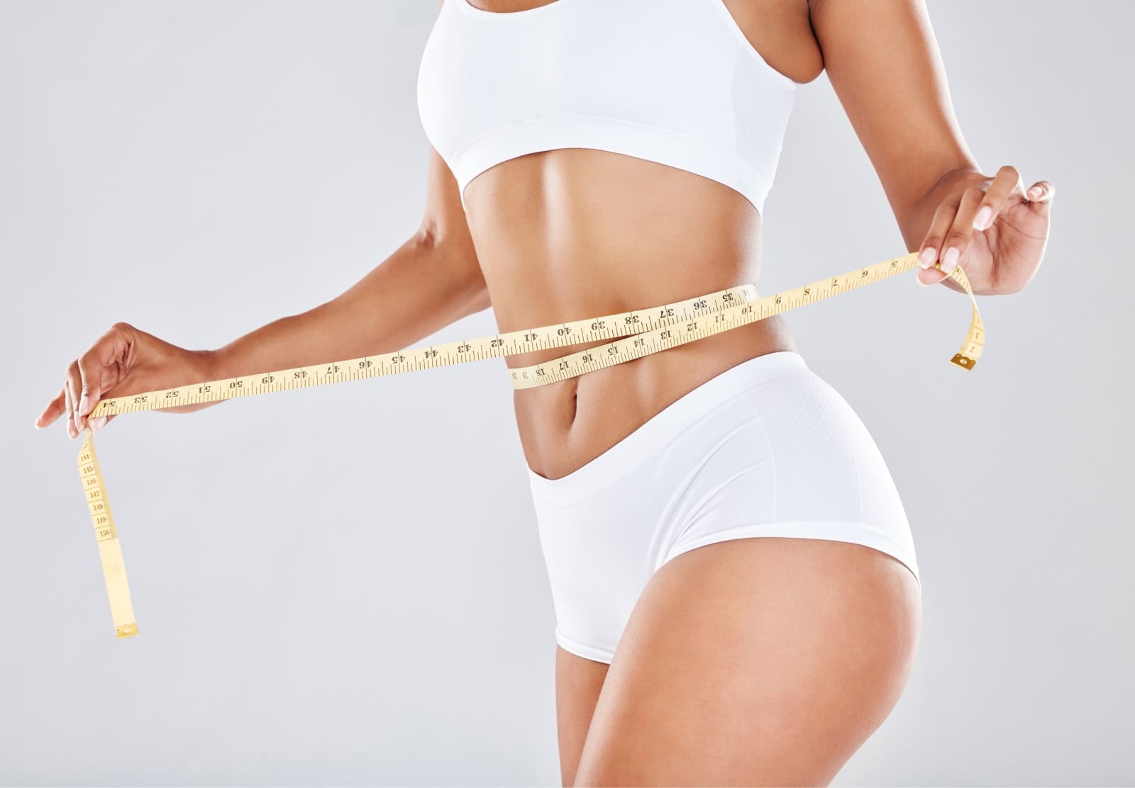 A close-up of a woman’s midsection wearing white activewear, holding a measuring tape around her waist, symbolizing the results of medical weight loss programs that promote healthy weight management and body confidence.